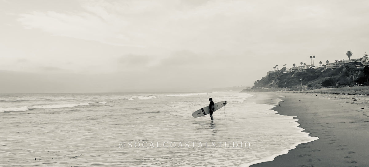 Black and white fine art print titled "The Walk In," showing a surfer walking into the water with footprints in the sand, from the Shoreline Editions collection by SoCal Coastal Studio.