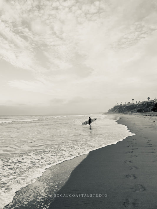 Black and white photo of a surfer with footprints in the sand along the San Clemente coast.